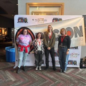Four people with red lanyards stand in front of a banner for the American CollegeTheater Festival inside a building with framed art on the walls.
