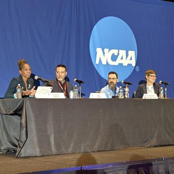 Four panelists sit at a table on stage with microphones, speaking in front of a blue backdrop displaying the NCAA logo.