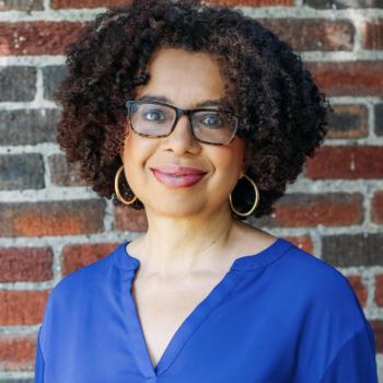 Woman with curly hair, glasses, and hoop earrings smiles at the camera, wearing a blue blouse and standing in front of a brick wall.
