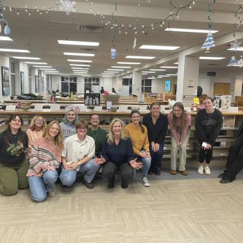 A group of students and a professor pose and smile for a photo in the Hilda C. Landers Library, with artist books and winter-themed decorations visible in the background.