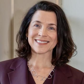 A woman with shoulder-length dark hair, wearing a maroon blazer and a silver chain necklace, smiles at the camera against a neutral background.