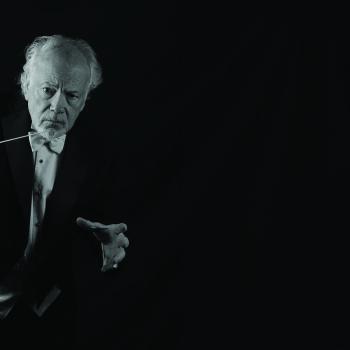An older man in formal attire holds a conductor’s baton and poses against a dark background, appearing to conduct music.