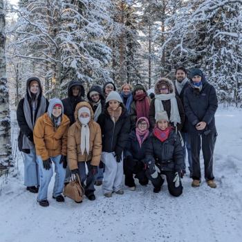 A group of fourteen people dressed in winter clothing pose for a photo on a snowy path surrounded by snow-covered trees.