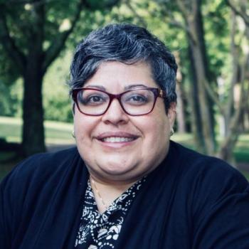 A person with short gray hair and glasses smiles outdoors, wearing a black and white patterned top and a black jacket. Trees and greenery are in the background.