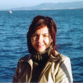 A woman with dark hair and a beige jacket stands in front of a large body of water with distant mountains in the background.