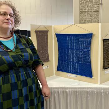 A woman stands next to a display featuring four geometric textile artworks and a tall, curved sculptural piece on a table.