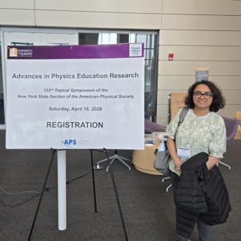 A woman stands next to a registration sign for the "Advances in Physics Education Research" symposium at the University at Albany, dated April 18, 2026.