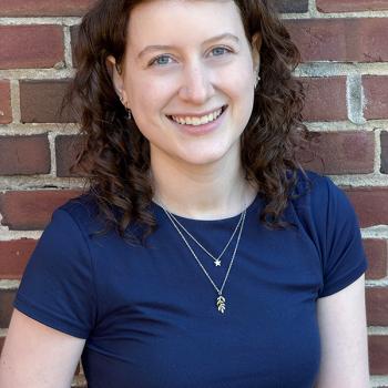 A woman with curly brown hair wearing a navy blue shirt and layered necklaces stands smiling in front of a brick wall.