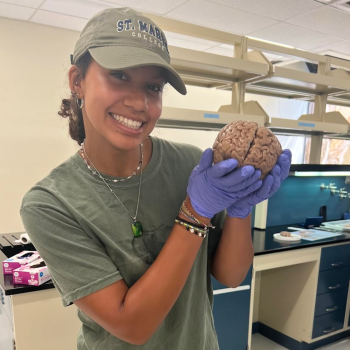 A woman wearing gloves and a cap holds a brain specimen in a laboratory setting, smiling at the camera.