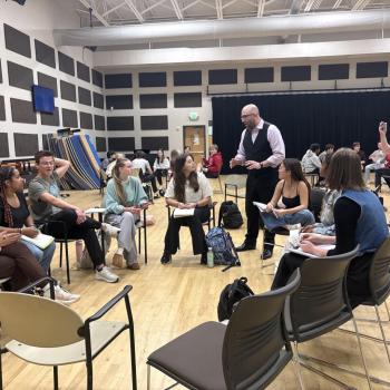 A group of students sits in a circle in a classroom while the instructor (Villada) stands and speaks; another person stands nearby and takes a photo with their phone.