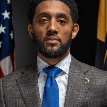 A man in a plaid suit jacket and blue tie stands in front of American and state flags, facing the camera with a neutral expression.