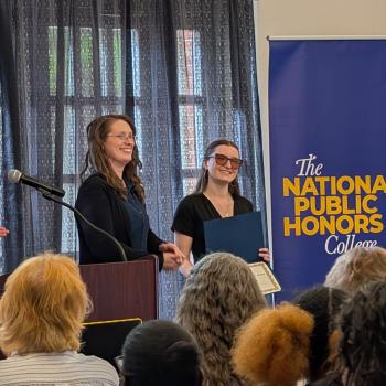 A woman receives an award on stage during a National Public Honors College event, while others applaud and a banner stands in the background.