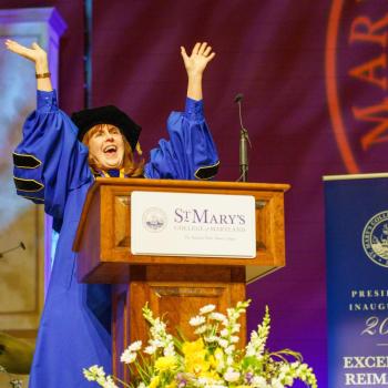 A person in academic regalia stands at a podium with arms raised, speaking at a St. Mary’s College of Maryland event, with a “Presidential Inauguration 2026” sign nearby.