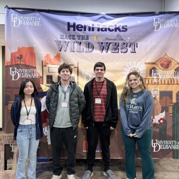 Four students stand in front of a "Hack the Wild West" HenHacks banner at the University of Delaware.