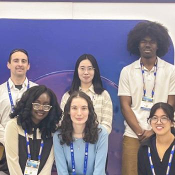 A group of eight people pose indoors in front of a colorful backdrop at what appears to be a conference or event, all wearing name badges.