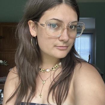A young woman with long brown hair wears clear glasses, hoop earrings, layered necklaces, and a plaid strapless top, standing indoors in natural light.