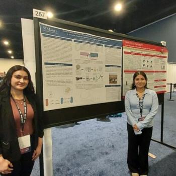 Two women stand beside a large scientific poster display at a conference with other posters and attendees visible in the background.