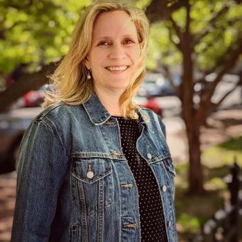 Woman with blonde hair wearing a denim jacket and a black, polka dot shirt stands outdoors in front of green trees, smiling at the camera.
