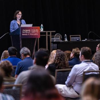 A woman speaks at a podium during a conference, addressing an audience seated in rows. A sign on the podium reads "ASBMB Make It Possible.