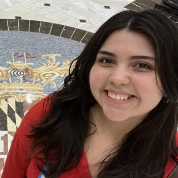 A young woman with long dark hair smiles at the camera, standing indoors on a marble floor featuring a decorative crest design.
