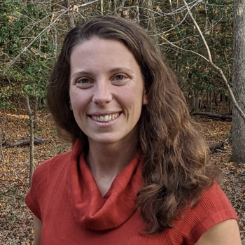 A woman with long brown hair, wearing a red cowl-neck top, stands outdoors in a wooded area with fallen leaves on the ground.