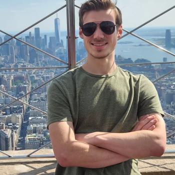 A man in sunglasses and a green shirt stands with arms crossed on an observation deck, with a city skyline and skyscrapers in the background.