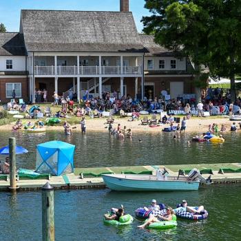 A crowd gathers at a lakeside beach near a large building, with people swimming, lounging on inflatables, and boats docked at a pier in the foreground.