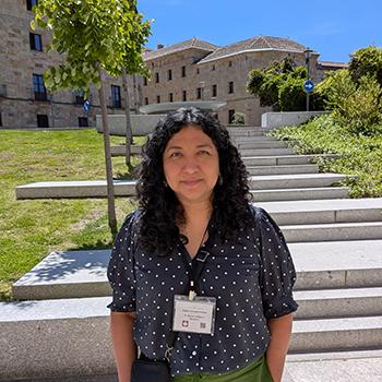 A woman with curly dark hair wearing a navy polka-dot blouse and name badge stands outdoors in front of stone steps and historic buildings on a sunny day.
