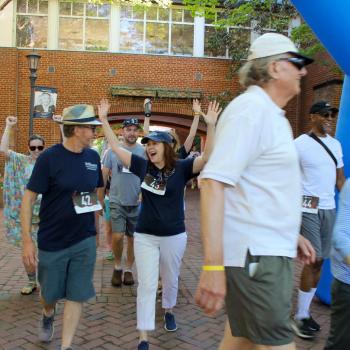 A group of people wearing numbered race bibs walk under a blue inflatable arch at an outdoor event; some are smiling and raising their arms.
