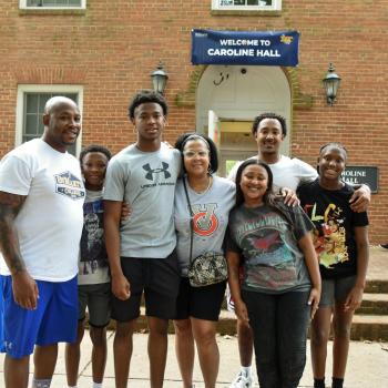 A group of six people stand and smile in front of a brick building with a “Welcome to Caroline Hall” sign above the door.