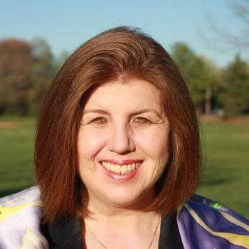 A woman with straight, shoulder-length brown hair smiles outdoors, wearing a black jacket and a colorful scarf, with trees and a blue sky in the background.