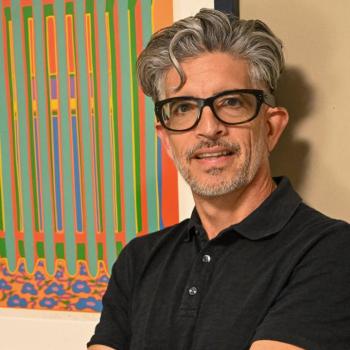 A man with gray hair, glasses, and a black polo shirt stands with arms crossed in front of a colorful abstract painting on a beige wall.