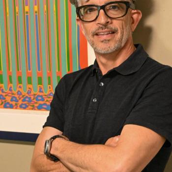 A man with gray hair, glasses, and a black polo shirt stands with arms crossed in front of a colorful abstract painting on a beige wall.
