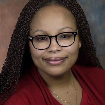 A woman with brown hair and glasses is wearing a red top, smiling at the camera against a neutral background.