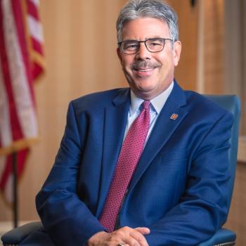 A man in a blue suit and red tie sits smiling in an office chair. An American flag and lamp are in the background.