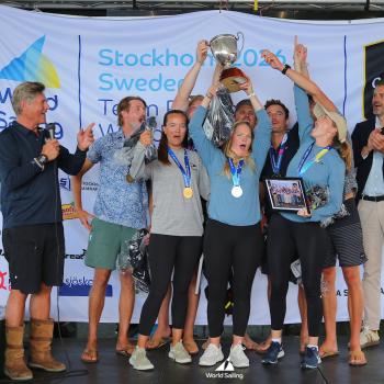 A group of people on stage celebrate with medals and a trophy at the World Sailing event, with banners and logos visible in the background. Photo by Malcom Hanes.