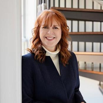 Woman with red hair wearing a navy blazer and white turtleneck stands smiling by a window, with shelves of binders in the background.