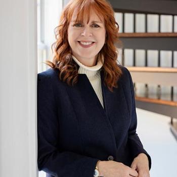 Woman with shoulder-length red hair in a navy blazer and white turtleneck stands indoors, smiling near a window with shelves of black binders in the background.