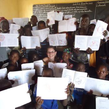 A group of children and an adult sit and stand in a classroom, holding up notebooks with drawings. A chalkboard with writing is visible in the background.