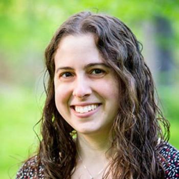 Woman with long curly brown hair smiles in an outdoor setting with a blurred green background.
