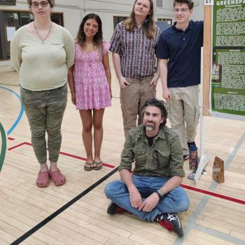 Five people pose by a science poster presentation in a gymnasium; four stand while one sits on the floor in front of the group.