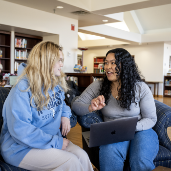 Two women sit on armchairs in a library, having a conversation. One holds a laptop on her lap, while bookshelves and another person are visible in the background.