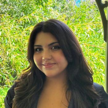 A woman with long dark hair and a black top stands in front of lush green foliage, smiling slightly at the camera.