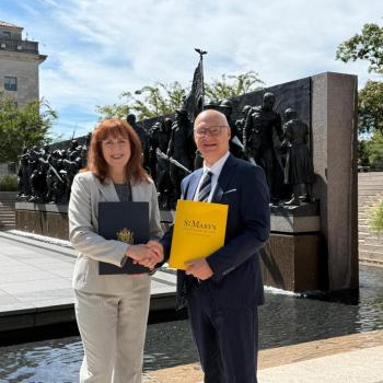 Two people in business attire shake hands outdoors in front of a large bronze monument, each holding official-looking folders.