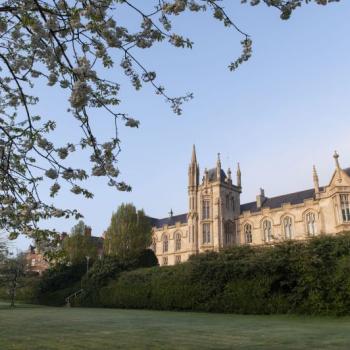 Large stone building with pointed towers sits behind a green lawn and blooming trees under a clear blue sky.
