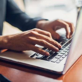 A person in business attire types on a laptop at a wooden desk, with part of a smartphone visible on the left side of the image.