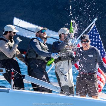 Four sailors celebrate on a sailboat, spraying champagne with an American flag in the background on a sunny day.
