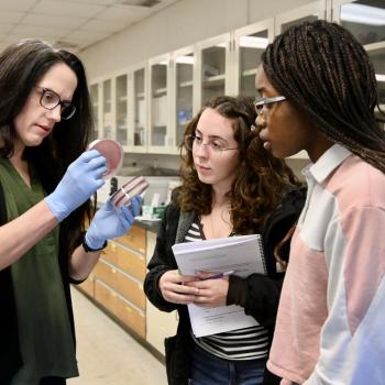 An instructor in a laboratory shows a petri dish to two students.