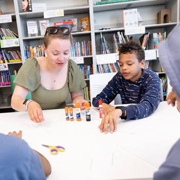 An adult and two children sit at a table working on a craft project with glue sticks and paper strips.