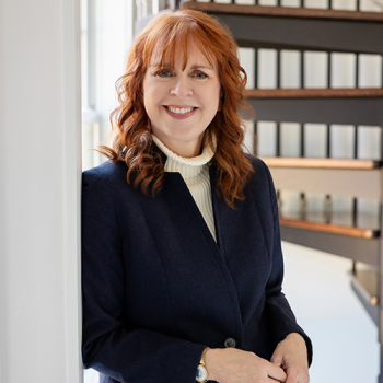 A smiling woman with shoulder-length auburn hair wearing a navy blue blazer over a cream-colored turtleneck.